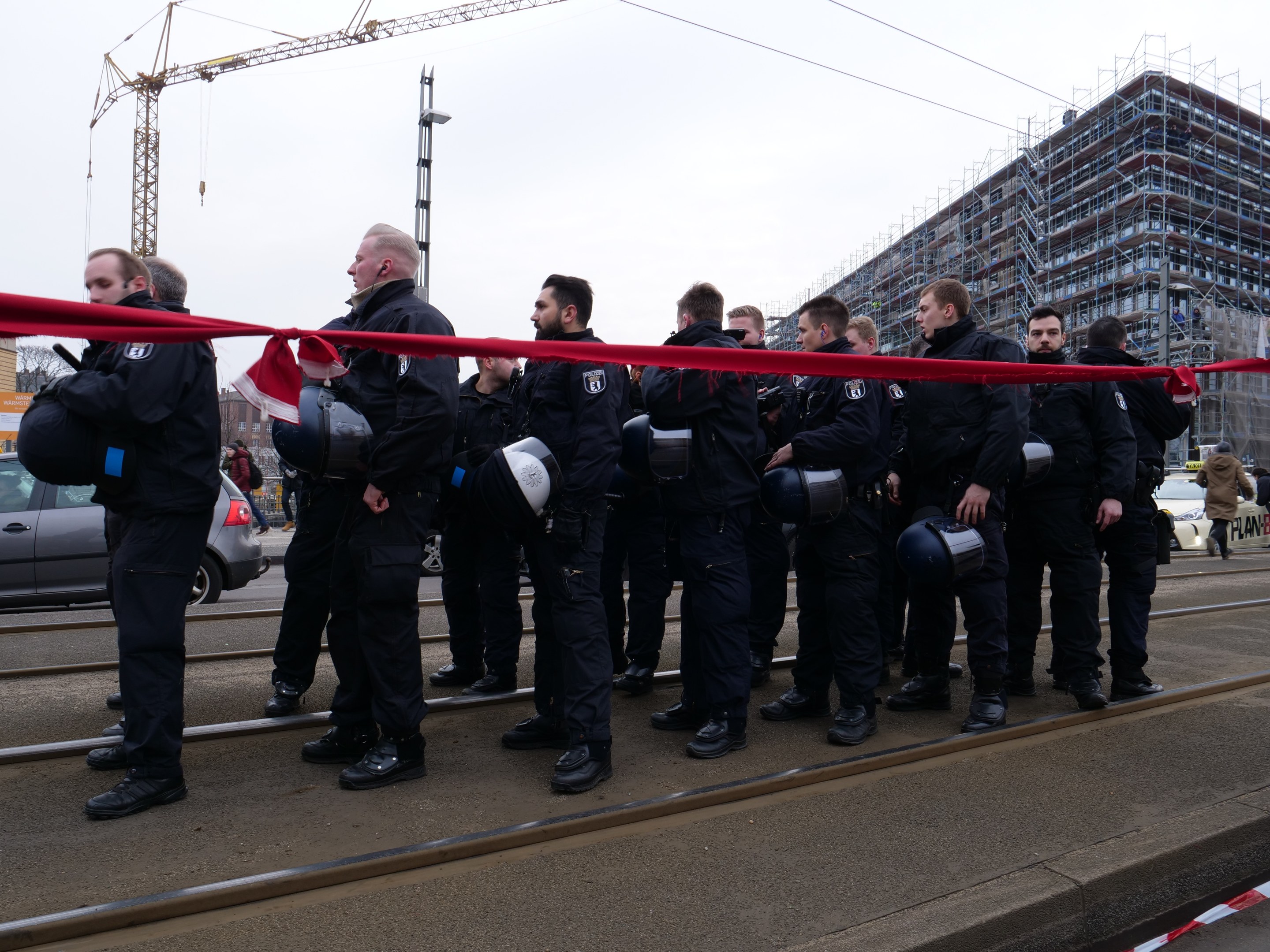 Eine Gruppe von Polizisten in schwarzen Uniformen und Helmen steht in einer Reihe auf einem Bahnsteig, mit einem roten Band davor, vor einer Kulisse aus Fahrzeugen, Passanten, Gebäuden und einem klaren blauen Himmel.