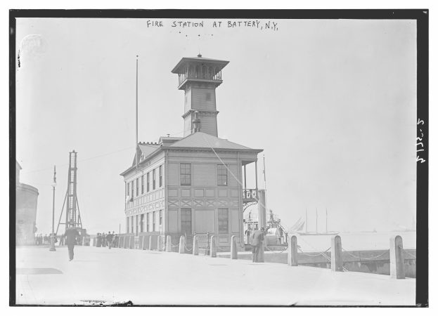 Ein Schwarz-Weiß-Foto einer Feuerwache an der Battery, New York, mit einem Gebäude, Menschen am Boden, Mästen, einem Zaun, Booten auf dem Wasser und einem bewölktem Himmel.