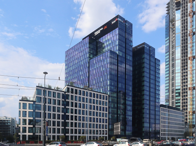 Großes Gebäude mit vielen Fenstern, die Deutsche Bank Hauptverwaltung in Frankfurt, Deutschland, umgeben von städtischen Elementen einschließlich Straßenlaternen, Fahrzeugen, Grünflächen und einem bewölkten Himmel.