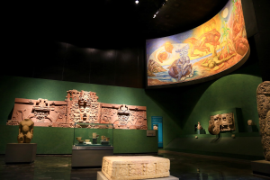 Ein Ausstellungsraum im Anthropologie-Museum von Mexiko-Stadt mit verschiedenen Artefakten, darunter Skulpturen, Tischgegenstände, eine Texttafel, eine Wandmalerei und Deckenbeleuchtung.