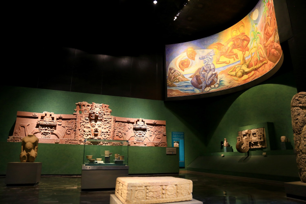 Ein Ausstellungsraum im Anthropologie-Museum von Mexiko-Stadt mit verschiedenen Artefakten, darunter Skulpturen, Tischgegenstände, eine Texttafel, eine Wandmalerei und Deckenbeleuchtung.