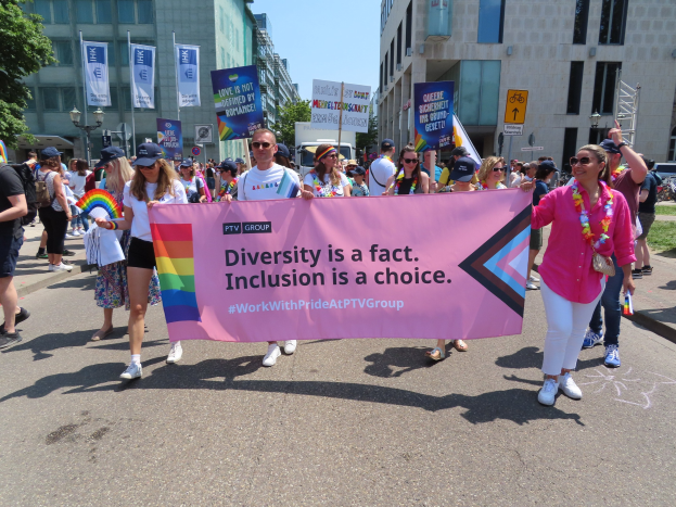 Eine Gruppe von Menschen marschiert auf einer Straße, hält ein Banner mit der Aufschrift "Diversität ist eine Tatsache, Inklusion ist eine Wahl" und trägt Kappen und Schutzbrillen, während andere zusätzliche Banner halten, vor einem Hintergrund aus Gebäuden, Bäumen, Laternen und einem klaren blauen Himmel.
