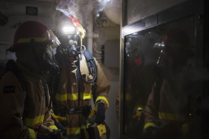 Feuerwehrleute in Gasmasken und Helmen vor einer raucherfüllten Tür in einem beengten Raum mit Rohren im Hintergrund.