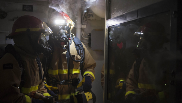 Feuerwehrleute in Gasmasken und Helmen vor einer raucherfüllten Tür in einem beengten Raum mit Rohren im Hintergrund.