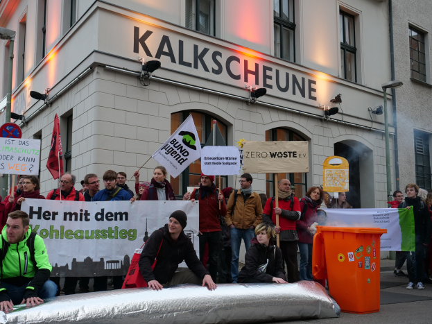 Eine Gruppe von Menschen demonstriert vor einem Gebäude in Deutschland und hält Schilder und Plakate hoch, während zwei Personen im Vordergrund sitzen und ein Müllcontainer rechts zu sehen ist.