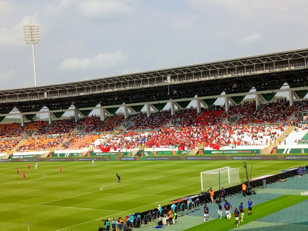 Ein Fußballspiel in einem großen Stadion mit Spielern auf dem Rasen, einem Tor, einem Zaun, einer Fahne, Metallrahmen, einem Dach mit Deckenbeleuchtung und einer bewölkten Himmel.