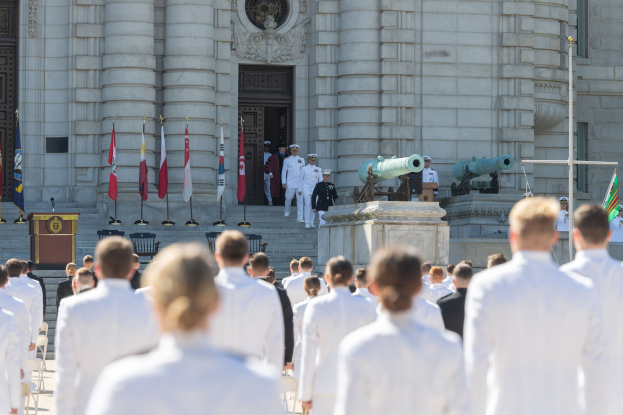 Gruppe von Menschen in weißen Marineuniformen vor einem neoklassischen Gebäude mit Säulen, einer Tür und Treppen, bei einer Abschlussfeier mit Fahnen, einem Podium, einem Mikrofon und Kanonen im Hintergrund.