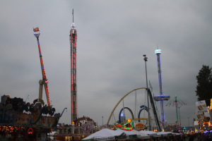 Eine Menschenmenge versammelt sich um ein hell erleuchtetes Faschingsfest in der Nacht, mit beleuchteten Fahrgeschäften, Pfählen und Bäumen unter einem bewölkten Himmel.