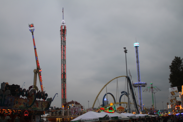 Eine Menschenmenge versammelt sich um ein hell erleuchtetes Faschingsfest in der Nacht, mit beleuchteten Fahrgeschäften, Pfählen und Bäumen unter einem bewölkten Himmel.