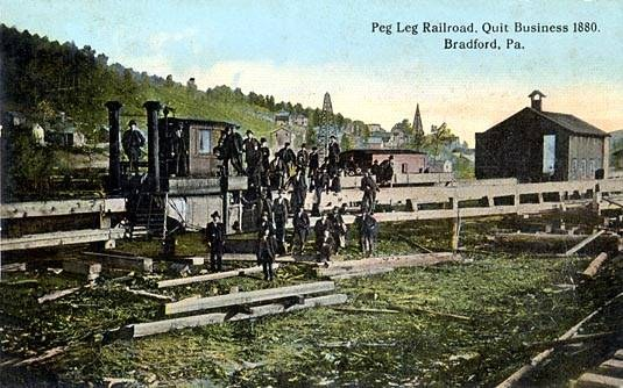 Gruppe von Menschen auf einem Bahngleis umgeben von einem Holzzaun, Häusern, Bäumen, Hügeln und einem klaren blauen Himmel mit der Aufschrift "Peg Leg Railroad, Quit Business 1880, Bradford, PA" oben.