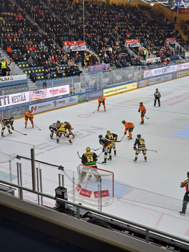 Eine Gruppe von Menschen, die Hockey auf einem Eisplatz spielen, umgeben von einem Zaun, mit Zuschauern im Hintergrund und beleuchteter Stadionbeleuchtung oben.