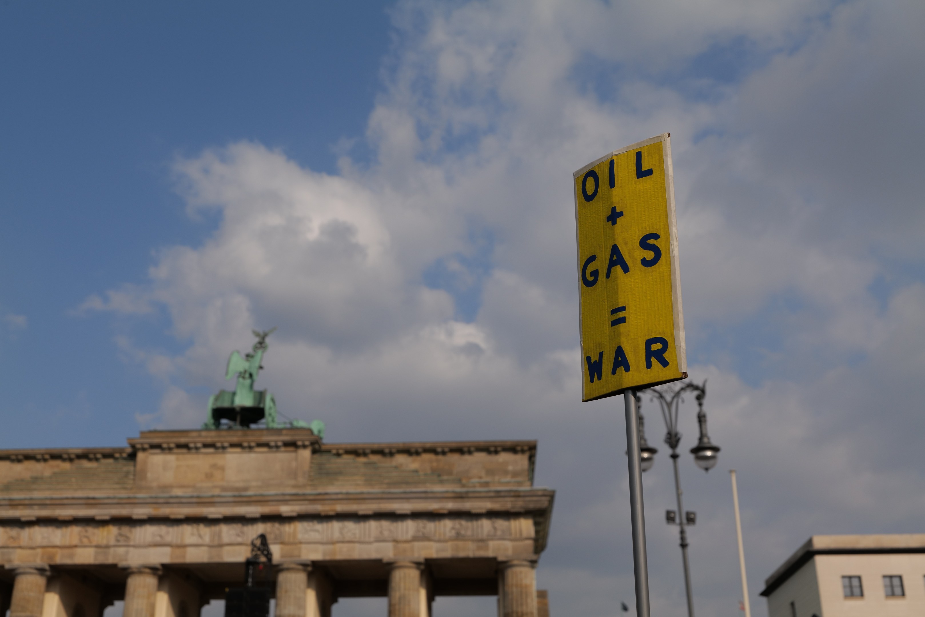 Gelbes Schild mit der Aufschrift "Oil and Gas War" im Vordergrund mit dem Brandenburg-Tor und Berliner Gebäuden im Hintergrund unter einem bewölktem Himmel.