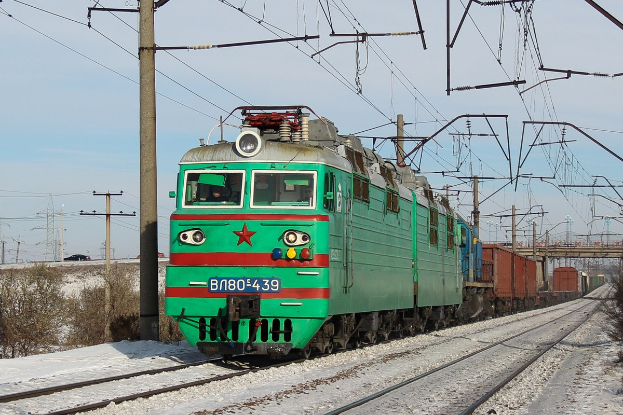 Ein grüner Elektrolokomotive fährt auf schneebedeckten Schienen, flankiert von Schnee und Pflanzen, mit Strommasten, Drähten, einer Brücke und einem klaren Himmel im Hintergrund.