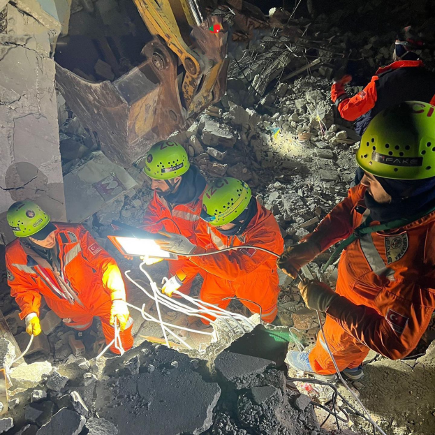 Gruppe von Menschen in orangefarbener Uniform und grünen Helmen bei der Arbeit an Schutt mit einem Kran im Hintergrund bei einem Einsatz nach dem Einsturz eines Gebäudes.