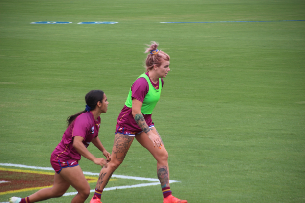 Zwei Frauen in Sportkleidung, eine in einem grünen T-Shirt und Shorts und die andere in einem pinken Kleid, spielen Fußball auf einem Feld, beide mit sichtbaren Tattoos an Händen und Füßen.