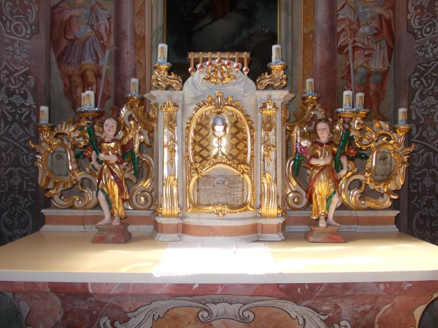 Ein Altar in einer Kirche mit Skulpturen von Menschen und brennenden Kerzen, vor einer Wand mit einem Gemälde, das eine feierliche Atmosphäre schafft.