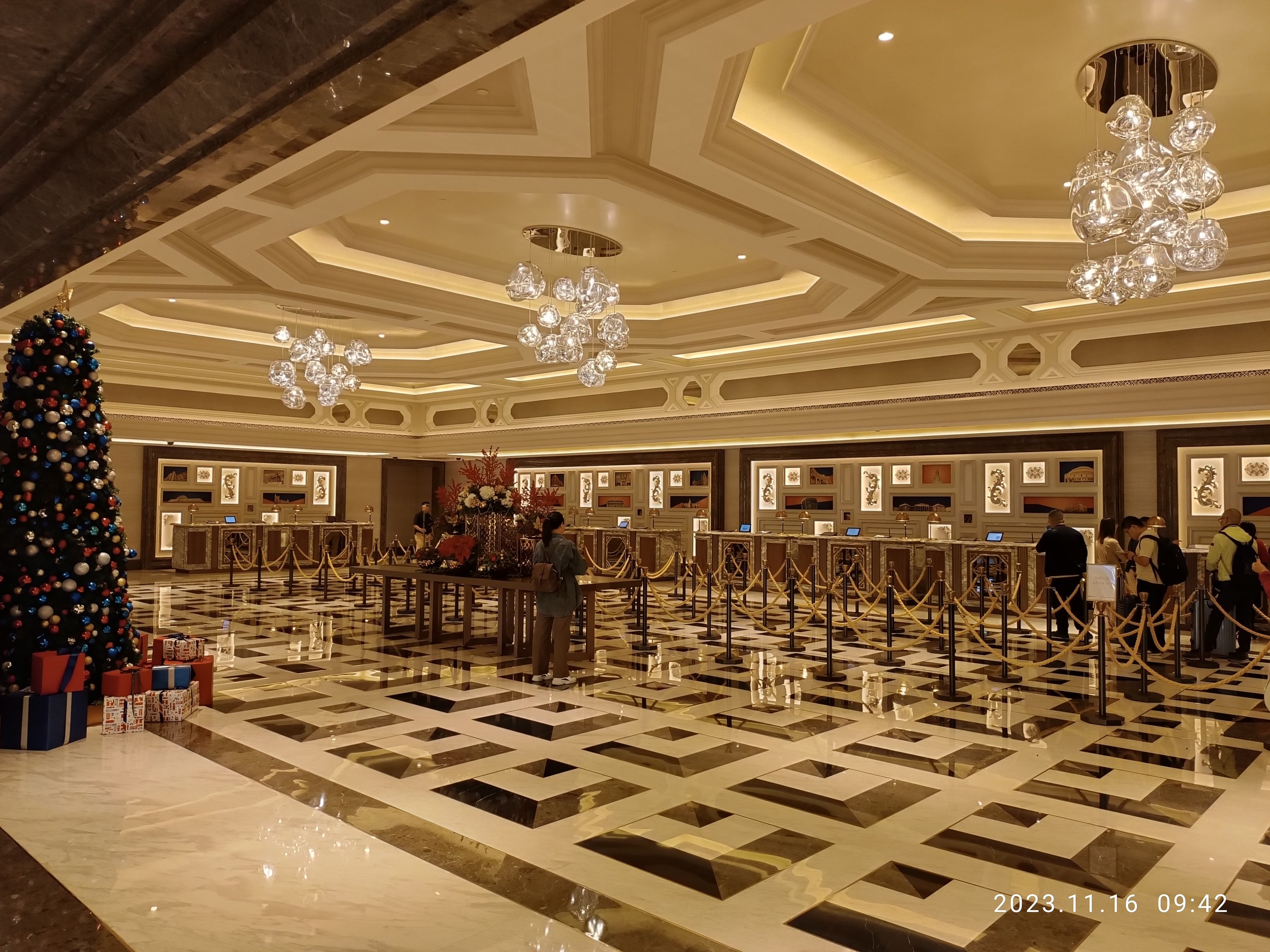 Ein geräumiger Eingangsbereich mit einem zentralen Weihnachtsbaum, umgeben von Geschenkboxen und festlichen Dekorationen, mit Menschen, Absperrpollern, Blumentischen, gerahmten Fotos an den Wänden und beleuchteter Decke.