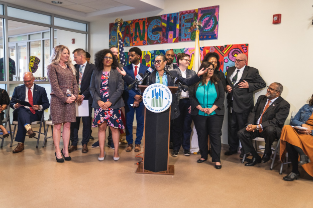 Gruppe von Menschen vor einem Rednerpult mit einem Mikrofon und einer Tafel mit der Aufschrift "Sancho Stadtrat tritt dem Stadtrat bei", mit sitzenden und stehenden Teilnehmern, Flaggen, Feuerlöscher und Deckenleuchten im Hintergrund.