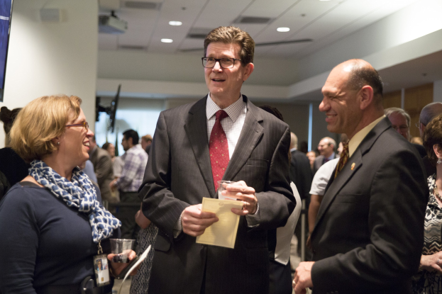 Eine Gruppe von Menschen in einem Raum, mit einem Mann in einem Anzug, der ein Papier und ein Glas in der Mitte hält, einer Frau in einem Schal, die links von ihm ein Telefon hält, einem Fernseher an der Wand und Deckenlampen darüber.