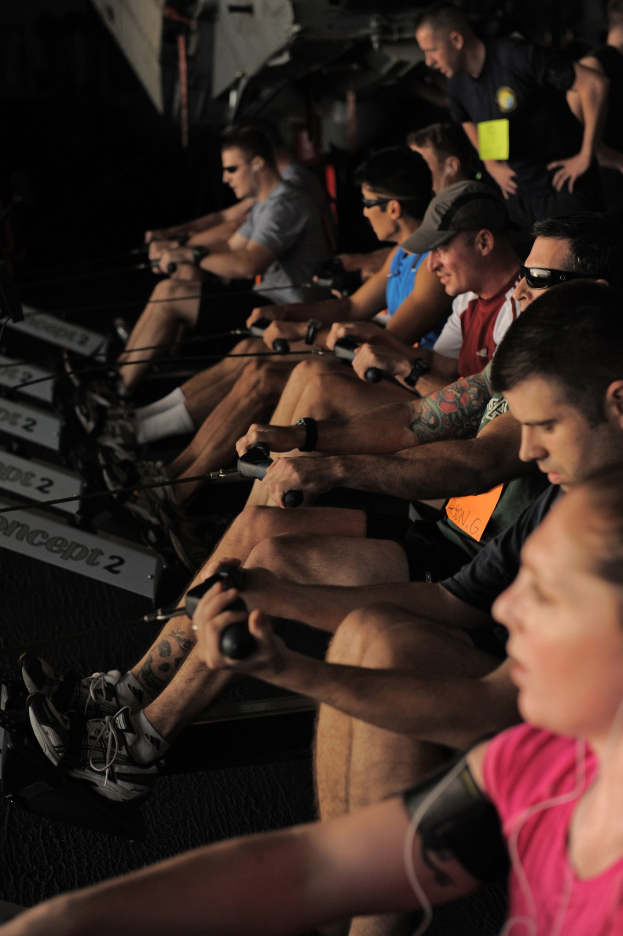 Eine Gruppe von Menschen, die Rudermaschinen in einem Fitnessstudio benutzen, mit anderen im Hintergrund, die alle Sportschuhe tragen.