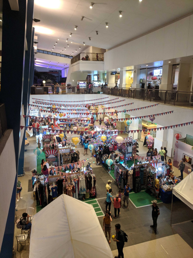 Ein volles Einkaufszentrum mit Ständen, hängenden Kleidern, Dekorationsflaggen, Ballons, sitzenden und stehenden Menschen, Geländern, Säulen und Deckenlampen, das an einen Weihnachtsmarkt im Mall of Asia erinnert.