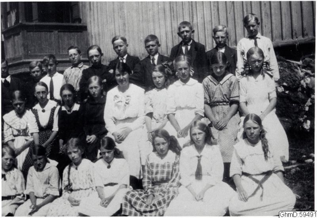 Schwarz-weißes Foto einer Gruppe von Kindern, die vor einem Gebäude mit Pflanzen im Hintergrund posieren, mit der Beschriftung "1910-1911 Schulkinder" unten.