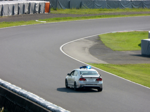 Polizeiauto auf einer Rennstrecke mit Gras auf beiden Seiten, Menschen, Zelte und Gegenstände im Hintergrund sichtbar.