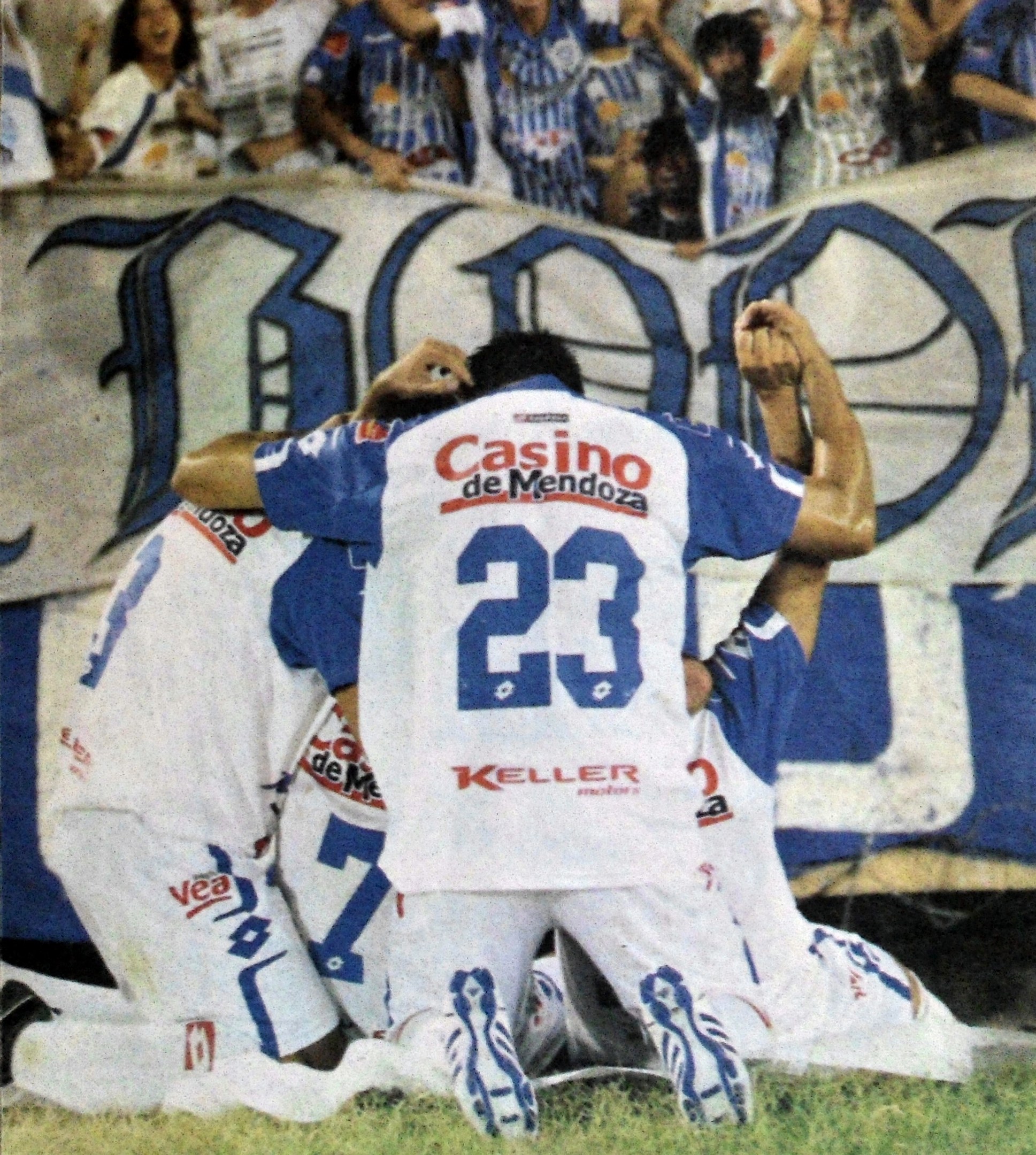 Gruppe von Fußballspielern feiert ein Tor auf dem Boden mit einer jubelnden Menge und einem Banner mit Text im Hintergrund.