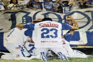 Gruppe von Fußballspielern feiert ein Tor auf dem Boden mit einer jubelnden Menge und einem Banner mit Text im Hintergrund.