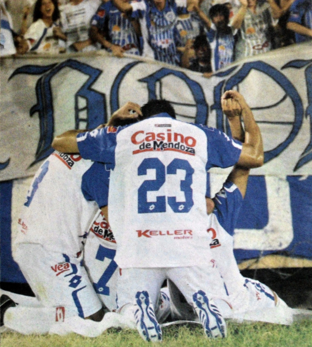 Gruppe von Fußballspielern feiert ein Tor auf dem Boden mit einer jubelnden Menge und einem Banner mit Text im Hintergrund.