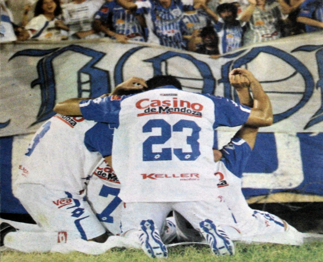 Gruppe von Fußballspielern feiert ein Tor auf dem Boden mit einer jubelnden Menge und einem Banner mit Text im Hintergrund.