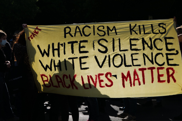 Eine Gruppe maskierter Personen auf einer dunklen Straße, die ein Banner halten, auf dem "Rassismus tötet, weiße Stille, weiße Gewalt, Black Lives Matter" steht.
