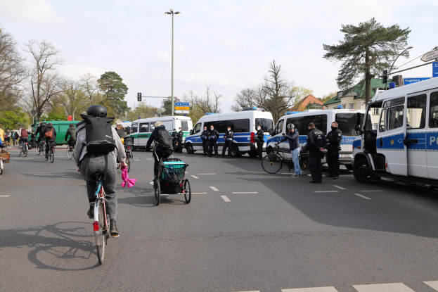 Gruppe von Menschen, die auf Fahrrädern eine Straße entlangfahren, mit Polizeiwagen, anderen Fahrzeugen, Bäumen, Laternen, Verkehrszeichen, Schildern, Gebäuden und einem bewölkten Himmel.