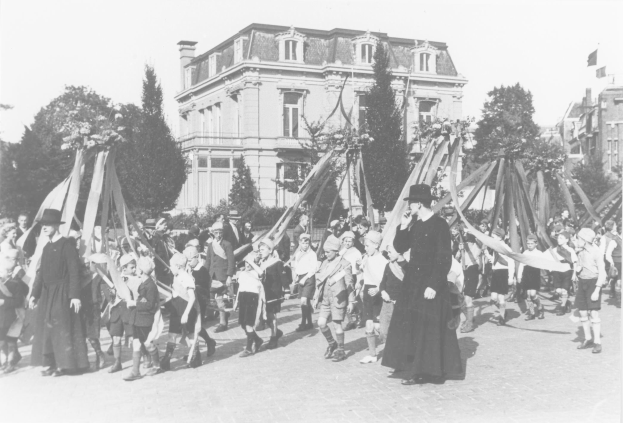 Ein Schwarz-Weiß-Bild einer Gruppe von Menschen, die eine Straße entlanggehen, einige halten Fahnen, mit Hüten, Bäumen, Gebäuden und einem sichtbaren Himmel im Hintergrund, das einen Maibaum als Symbol für den Maifeiertag zeigt.