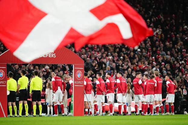 Menschen auf einem Fußballfeld mit einer roten und weißen Flagge im Vordergrund, einem Bogen mit der Aufschrift "Bayern München vs. Bayern München Wetten & Vorschau" im Hintergrund und einer großen Menge im Stadion.