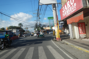 Eine belebte Stadtstraße mit Fahrzeugen, Fußgängern, Strommasten, Gebäuden, Bäumen und einem bewölkten Himmel, mit einem Pfandhaus namens "Jaro Pfandhaus" auf der rechten Seite.