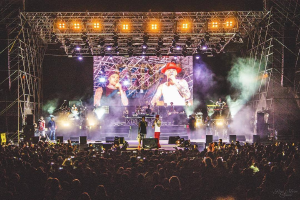 Eine Gruppe von Menschen auf einer hell erleuchteten Bühne in der O2 Arena London, einige spielen Instrumente und andere halten Telefone hoch, vor dunklem Hintergrund mit einem Live-Aufnahme-Wasserzeichen in der rechten unteren Ecke.