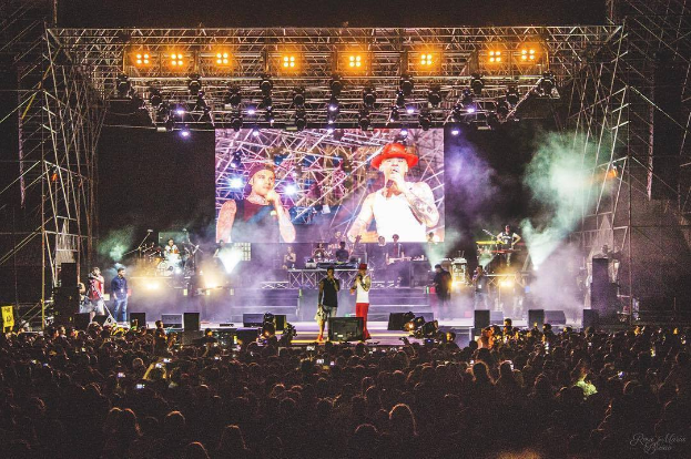 Eine Gruppe von Menschen auf einer hell erleuchteten Bühne in der O2 Arena London, einige spielen Instrumente und andere halten Telefone hoch, vor dunklem Hintergrund mit einem Live-Aufnahme-Wasserzeichen in der rechten unteren Ecke.
