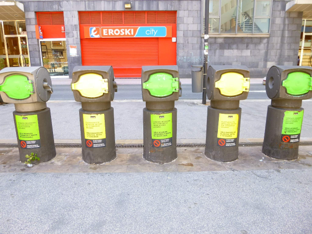 Eine Reihe von Parkscheinautomaten entlang einer Straße mit einem Gebäude mit Glasfenstern und einer Rolllade mit Text sowie einem nahen Müllcontainer.