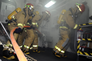 Feuerwehrleute in Schutzausrüstung mit Helmen und Sauerstoffflaschen arbeiten an einem Feuerlöschfahrzeug, mit einem Gestell voller Gegenstände und Hintergrundausrüstung im Hintergrund.