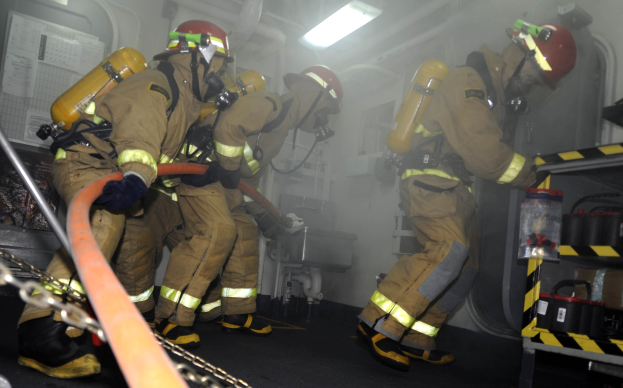 Feuerwehrleute in Schutzausrüstung mit Helmen und Sauerstoffflaschen arbeiten an einem Feuerlöschfahrzeug, mit einem Gestell voller Gegenstände und Hintergrundausrüstung im Hintergrund.