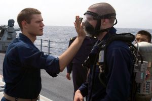 Ein Mann in einer Gasmaske und einem blauen Kleid steht rechts auf dem Deck eines Schiffs und spricht zu einem anderen Mann vor ihm, mit dem Ozean und dem Himmel im Hintergrund.