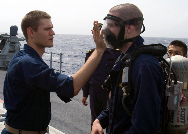 Ein Mann in einer Gasmaske und einem blauen Kleid steht rechts auf dem Deck eines Schiffs und spricht zu einem anderen Mann vor ihm, mit dem Ozean und dem Himmel im Hintergrund.