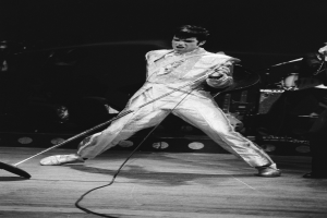 Elvis Presley performs on stage in a white jumpsuit, holding a microphone, with another musician to his right and stage lights and instruments in the background.