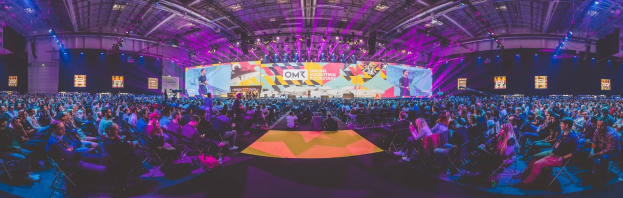 Eine große Zuschauermenge sitzt einem hell erleuchteten Bühne mit Bildschirmen gegenüber, wo mehrere Personen stehen während eines Online-Marketing-Events.