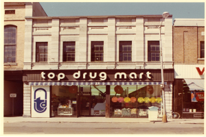 Top-Drogenmart-Filiale an einer Straßenecke mit Fenstern, einem Schild, einem Straßenschild, einem geparkten Fahrrad, einem Müllcontainer und einem bewölkten Himmel.