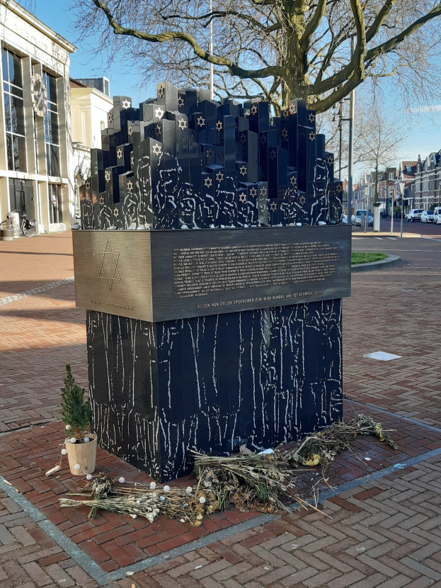 Denkmal in einem Stadtplatz für Holocaust-Opfer mit einem Baum davor, einem Topfpflanze auf dem Boden, verstreuten trockenen Blättern und umliegenden Gebäuden, Fahrzeugen, Polen und Bäumen.