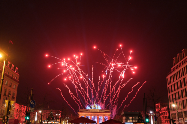 Eine Straßenansicht am Silvesterabend in Berlin mit Gebäuden, Bäumen, Laternenmasten, Verkehrszeichen, Schildern, Zelten, Menschen und einem Feuerwerk am Himmel.