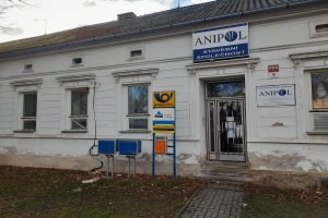 Weißer Bau mit einer Tafel, die "Anipol Post Office" zeigt, mit Fenstern, einer Tür, einer Tafel mit Text, Pfosten, einem Weg, Gras, trockenen Blättern, einem Baum und einem bewölkten Himmel.