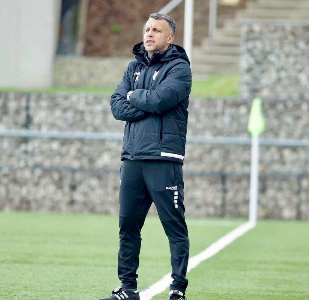 Ein Mann in schwarzer Jacke und Hose steht auf einem Fussballfeld mit überkreuzten Armen, der als Trainer während einer Trainingseinheit erscheint, mit einem Zaun, Treppe und Wand im Hintergrund.